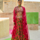 Woman in a vibrant traditional outfit with pink and green patterns standing in front of a stone wall.