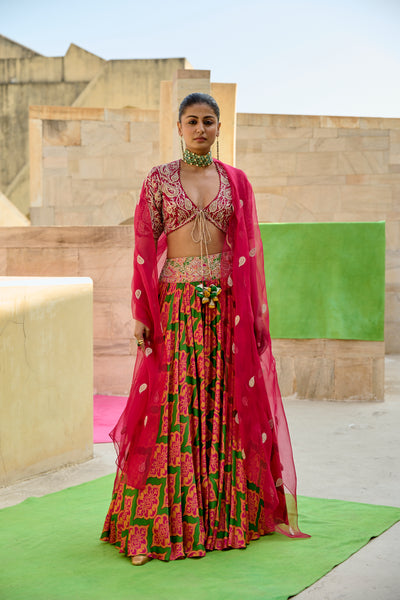 Woman in a vibrant traditional outfit with pink and green patterns standing in front of a stone wall.