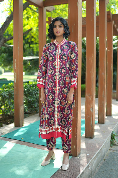 Woman wearing a patterned outfit standing in front of wooden pillars with greenery in the background
