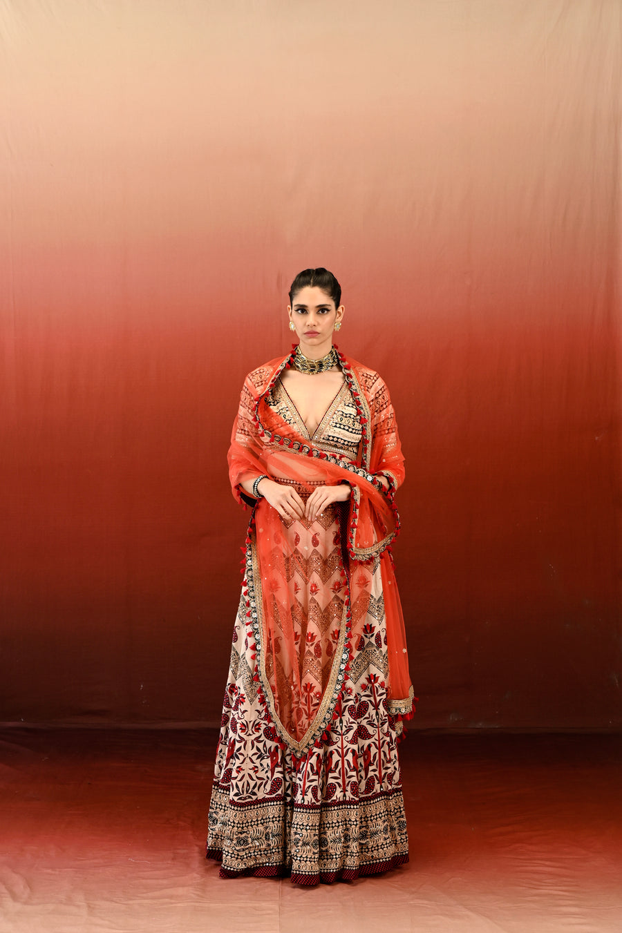 A woman standing in a red background, wearing a traditional Paisley Lehenga Set with a V-neck blouse and a dupatta.