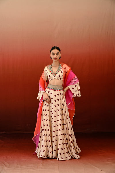 A woman standing in a red background wearing a traditional ecru and rust lehenga set with hand embroidery, including a V-neck blouse and a dupatta.