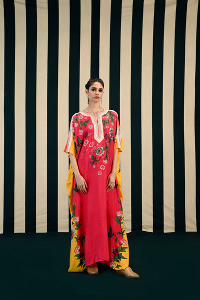 A woman standing against a striped background, wearing a fuchsia-colored kaftan with floral embroidery and a side slit, paired with a yellow dupatta with floral patterns.
