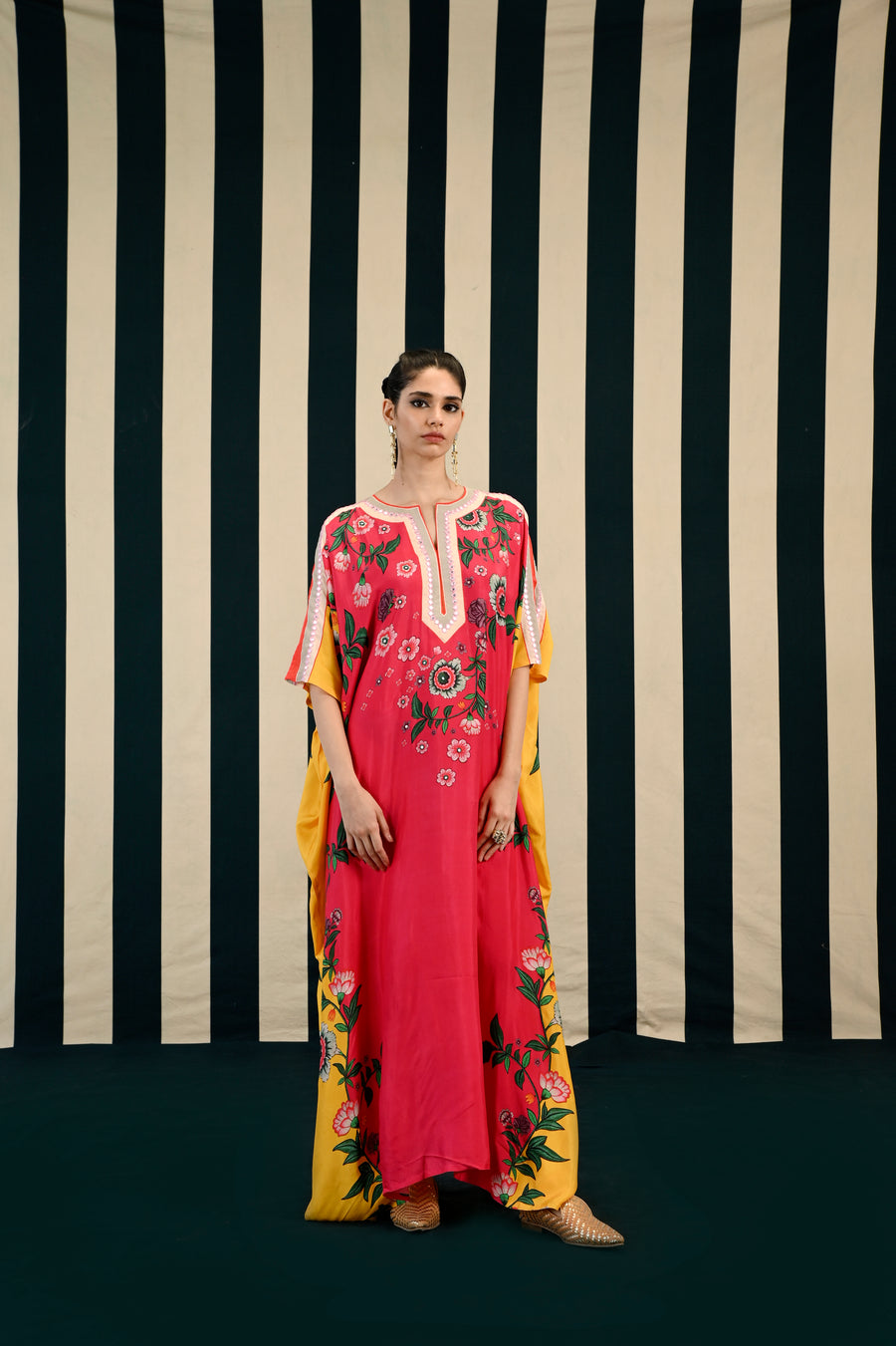 A woman standing against a striped background, wearing a fuchsia-colored kaftan with floral embroidery and a side slit, paired with a yellow dupatta with floral patterns.