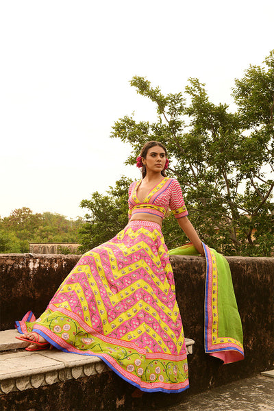 A woman sitting on a stone bench wearing a traditional, multicolored lehenga set with floral and geometric patterns, paired with a dupatta.