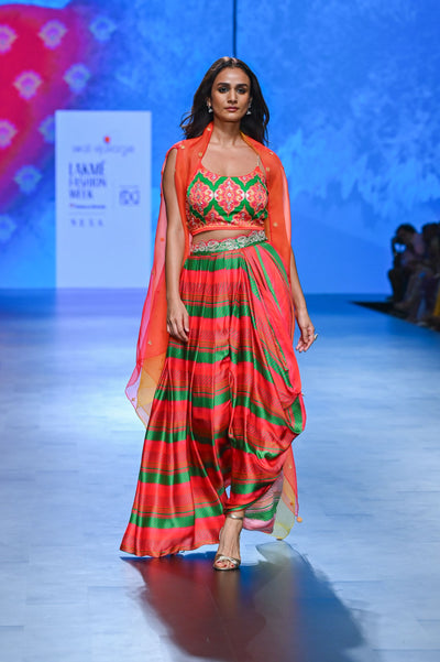 A model walking on a ramp showcasing a red and emerald green striped organza cape with a singlet and dhoti, at a fashion week.